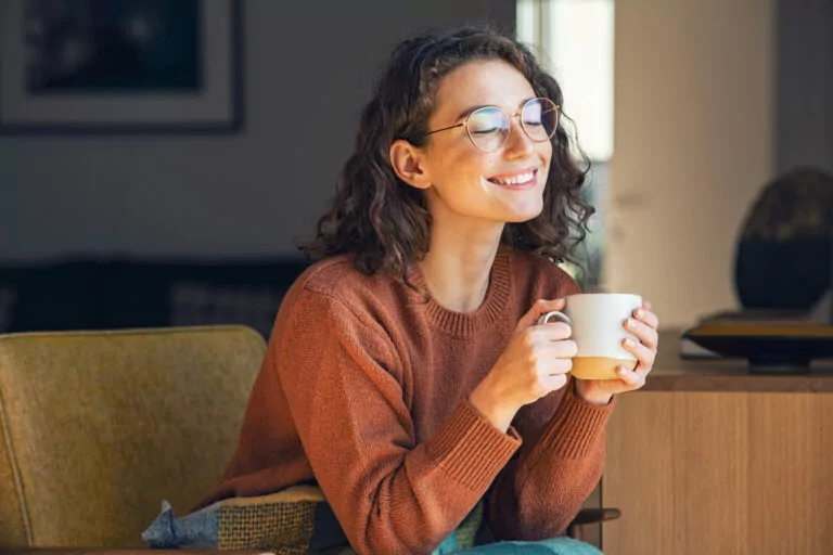 A young woman with curly hair and glasses sits comfortably, holding a mug with both hands. She is smiling with her eyes closed, wearing a rust-colored sweater, and appears content and relaxed in a cozy indoor setting.