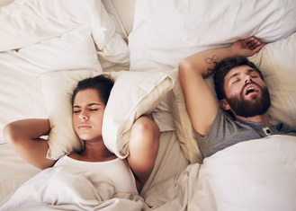 Two people lying in bed, one covering ears with pillow while other sleeps on back with mouth open, suggesting snoring disturbance.