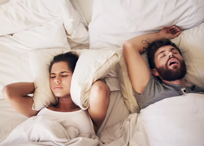 A couple is lying in bed with white sheets and pillows. The man on the right is sleeping with his mouth open and arms above his head. The woman on the left appears frustrated, covering her ears with a pillow to block out the noise. She has a pained expression on her face.