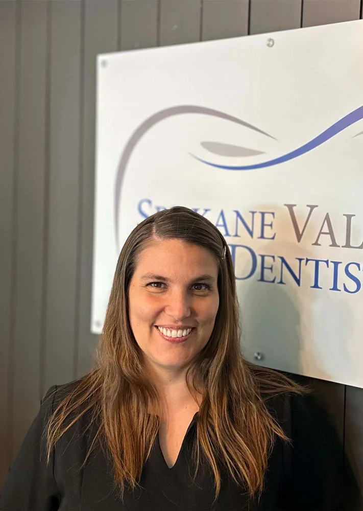 Headshot of Amanda wearing black scrubs, standing in front of the logo of "Spokane Valley Dentistry."