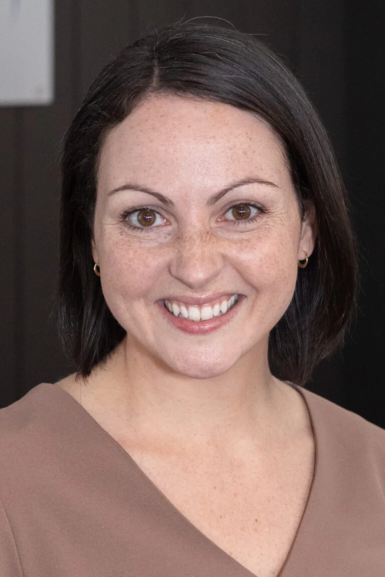 Person with short, dark hair wearing a brown V-neck top, shown against a plain background.