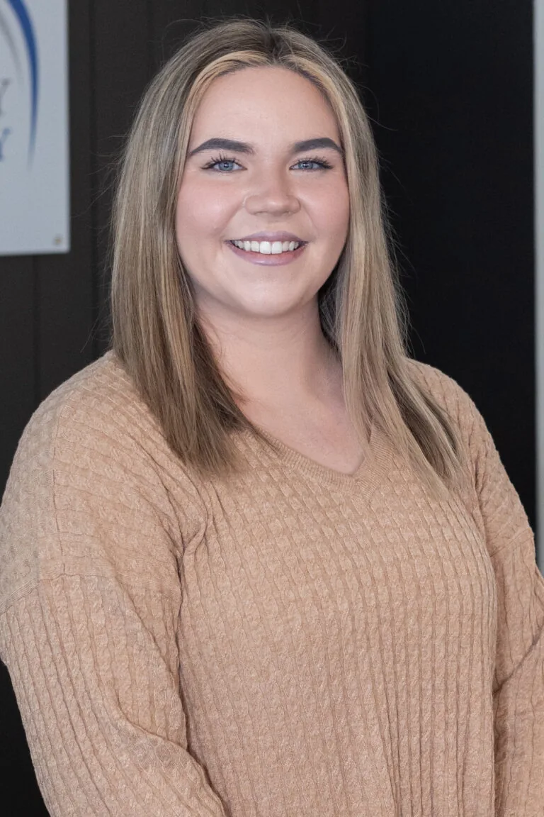 Person wearing a ribbed, light brown sweater with straight, highlighted hair against a dark background, partially showing a sign on the left.
