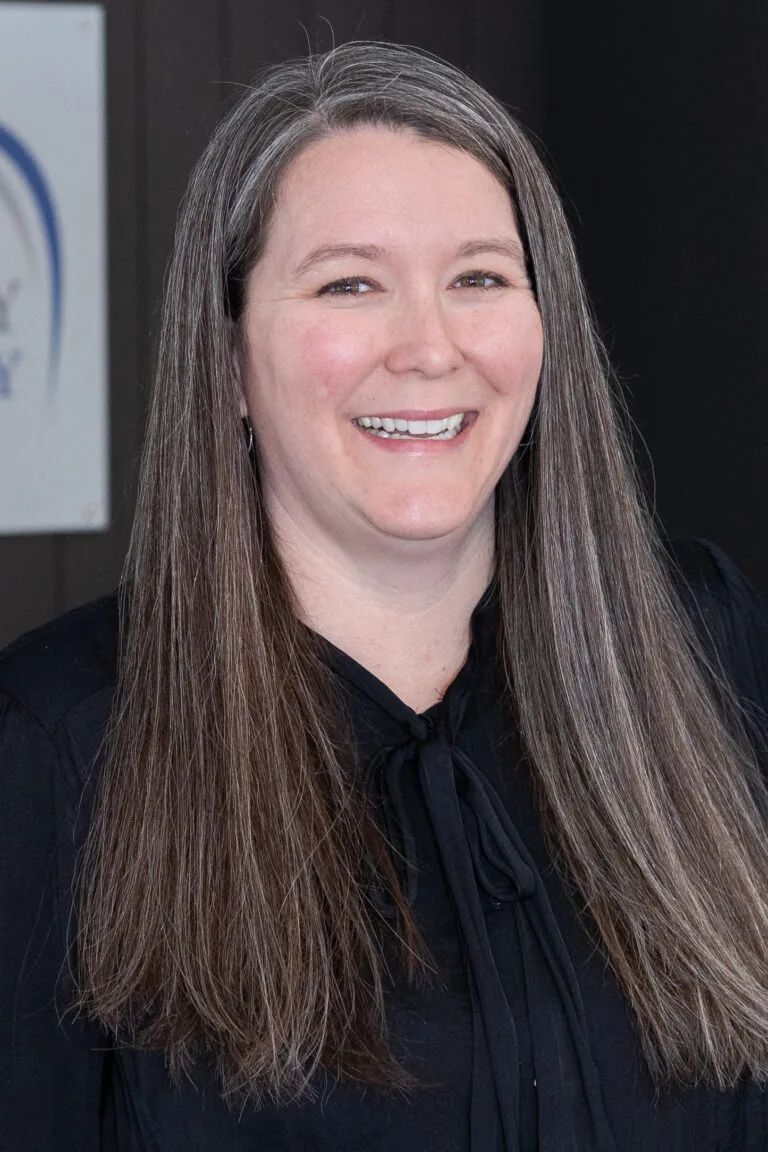Individual with long, straight hair that is a mix of gray and brown, wearing a black top with a tied bow. The part of a sign with a blue logo is visible in the background.