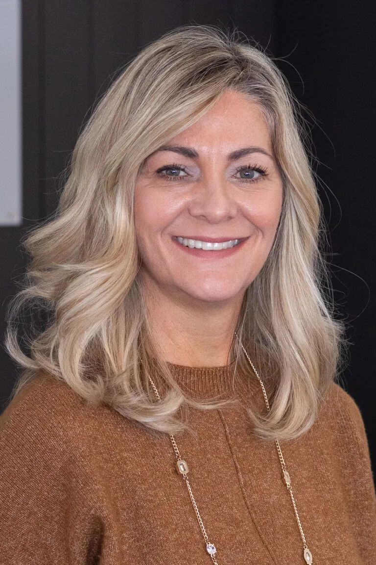 A person with shoulder-length blonde hair, wearing a brown sweater and a beaded necklace.